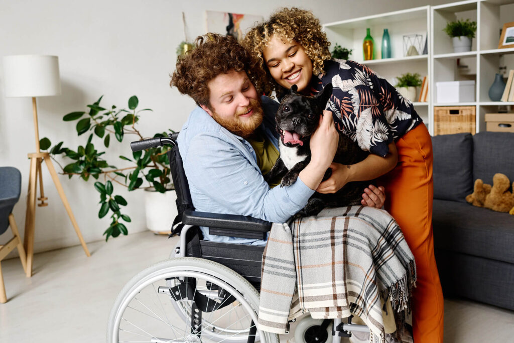 Couple embracing a dog who is sitting on the lap of a man in a wheelchair
